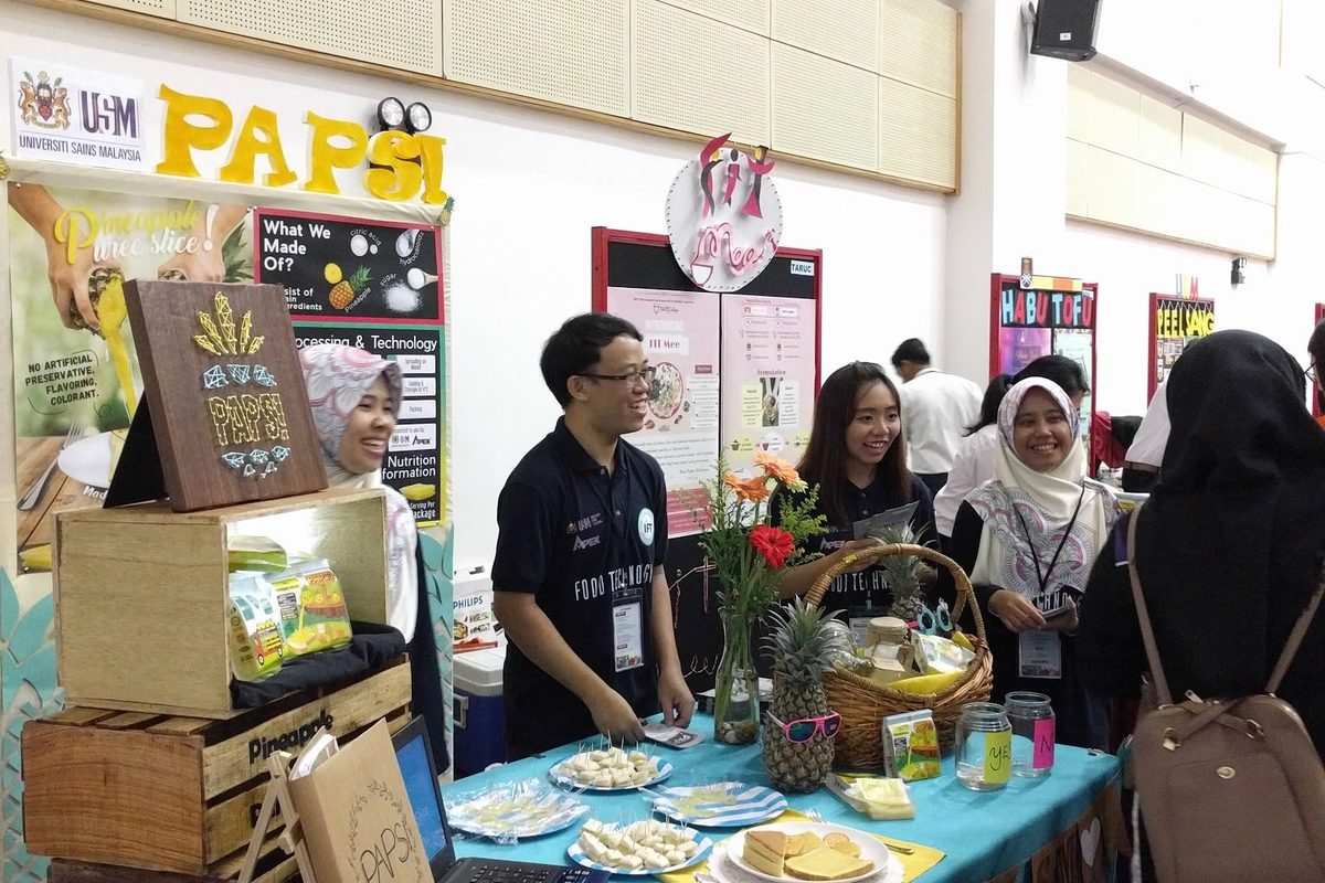 Students at a decorated food product booth talk with visitors in the hall