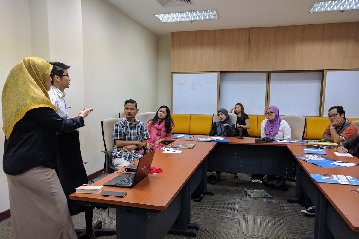 Two speakers stand at the front of a meeting room while participants at a U-shaped table listen