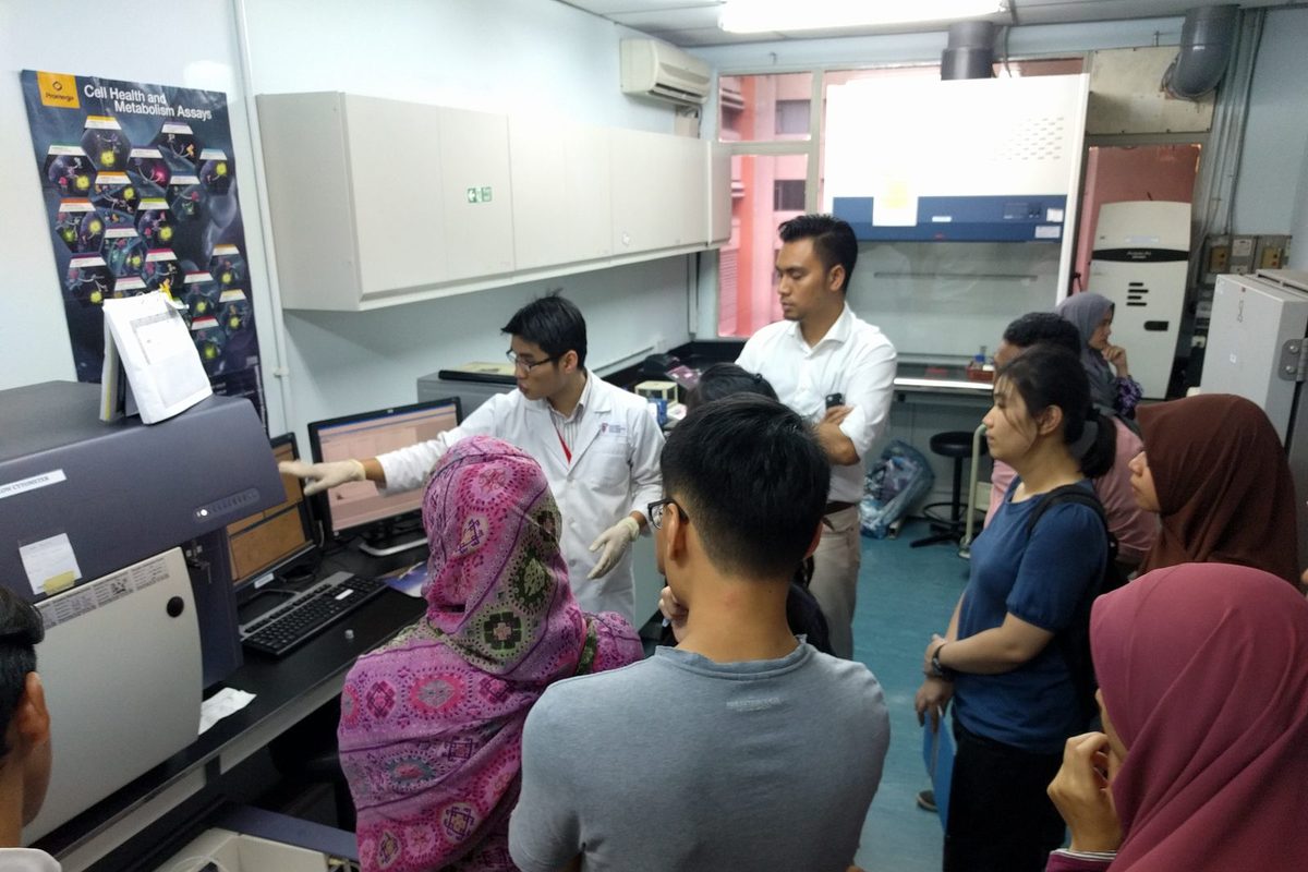 A presenter in a lab coat points to the flow cytometer screen while participants watch