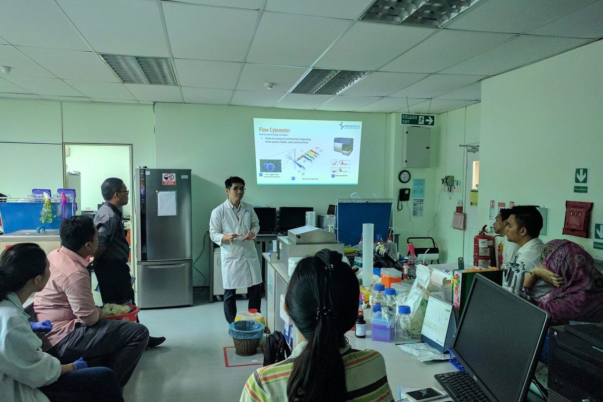 A presenter in a lab coat gives a talk beside a projected slide titled Flow Cytometer