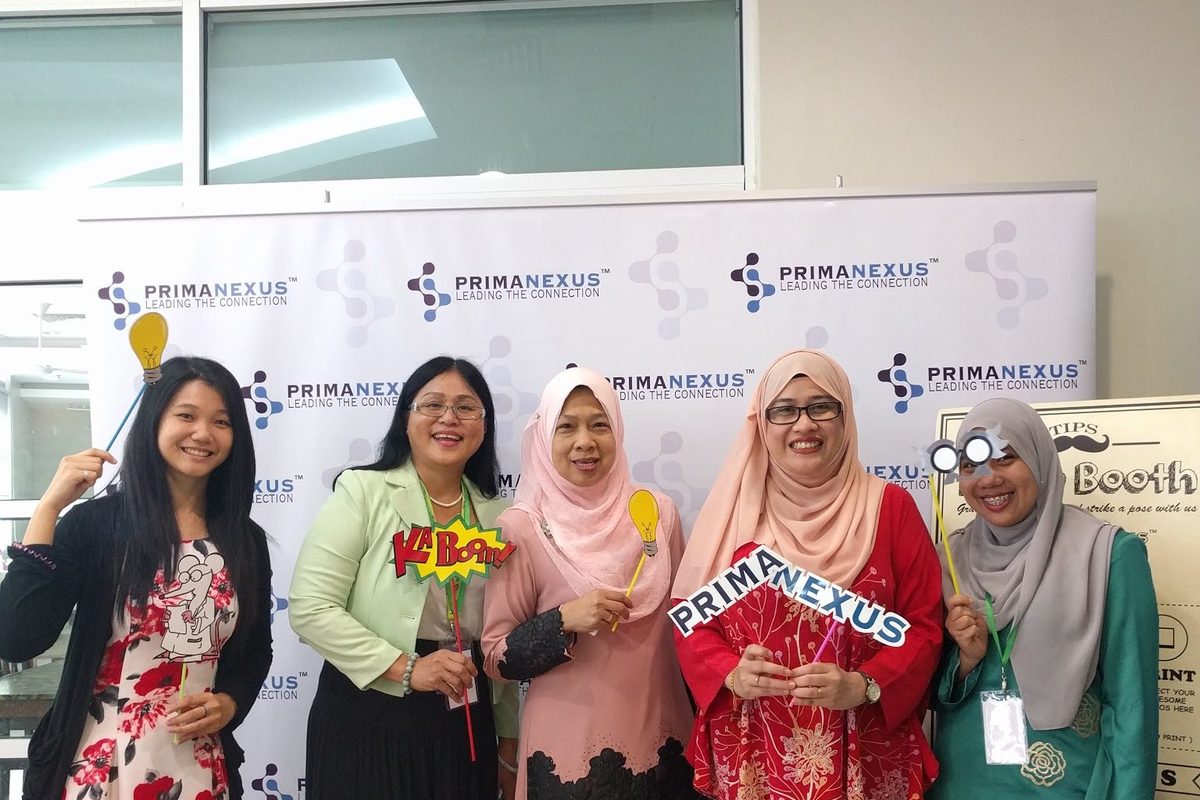 Five women hold photo props in front of the Prima Nexus backdrop at the photo booth