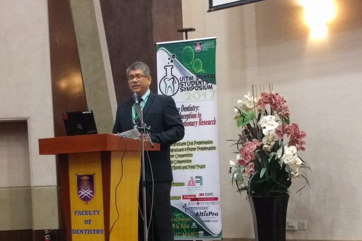 A speaker at the lectern on the symposium stage, beside the event banner and a flower arrangement