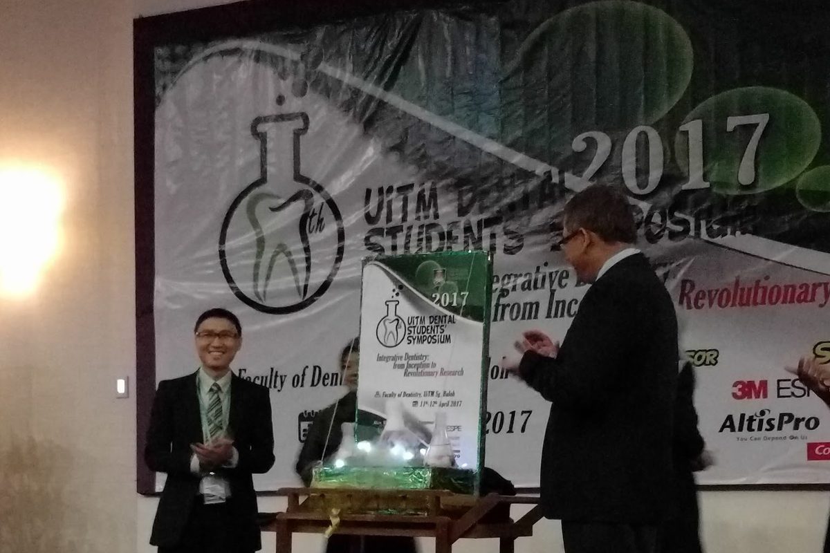 Two men in suits on stage beside a glass display on a trolley, in front of the symposium backdrop