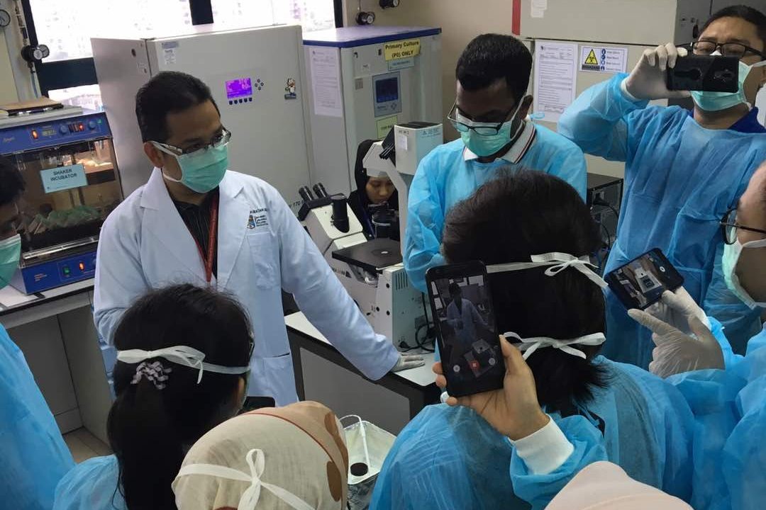 A demonstrator in a white coat at a lab bench while participants in gowns record on their phones