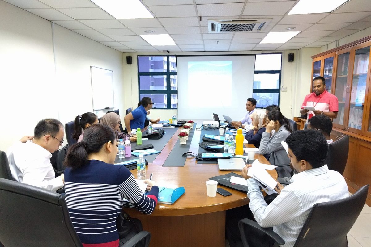Participants seated around a long table in a meeting room, with a presentation on the screen
