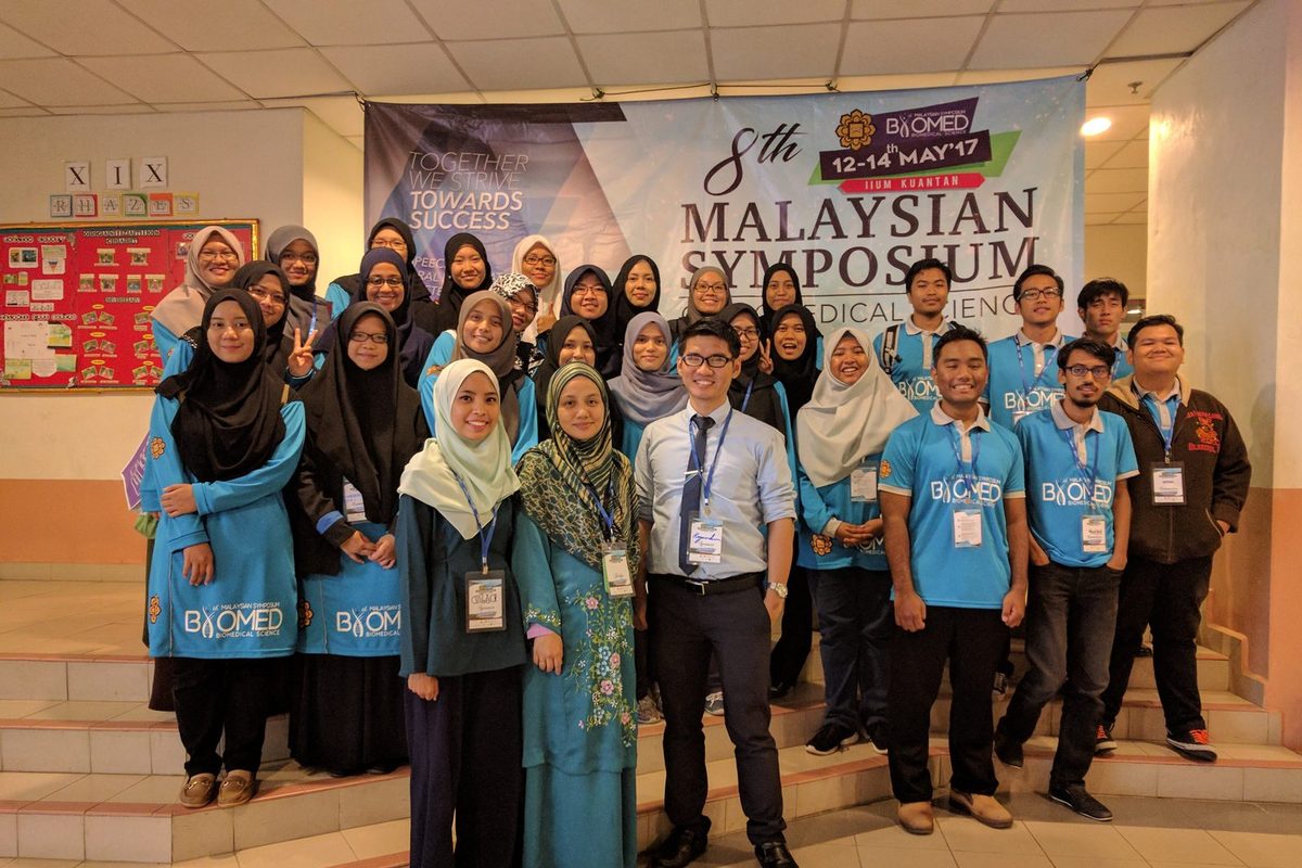 A large group, many in blue symposium T-shirts, poses in front of the 8th Malaysian Symposium of Biomedical Sciences banner
