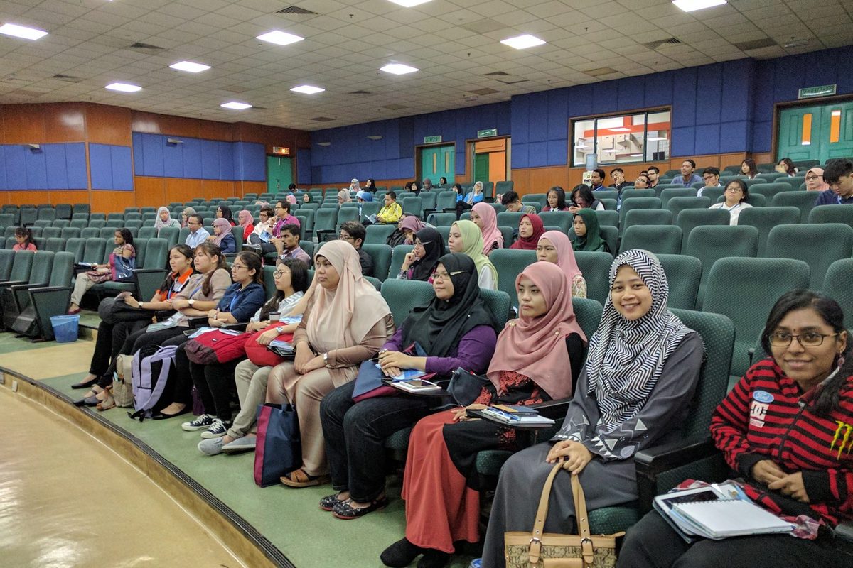 Participants seated in rows in a lecture hall, some with notes on their laps.