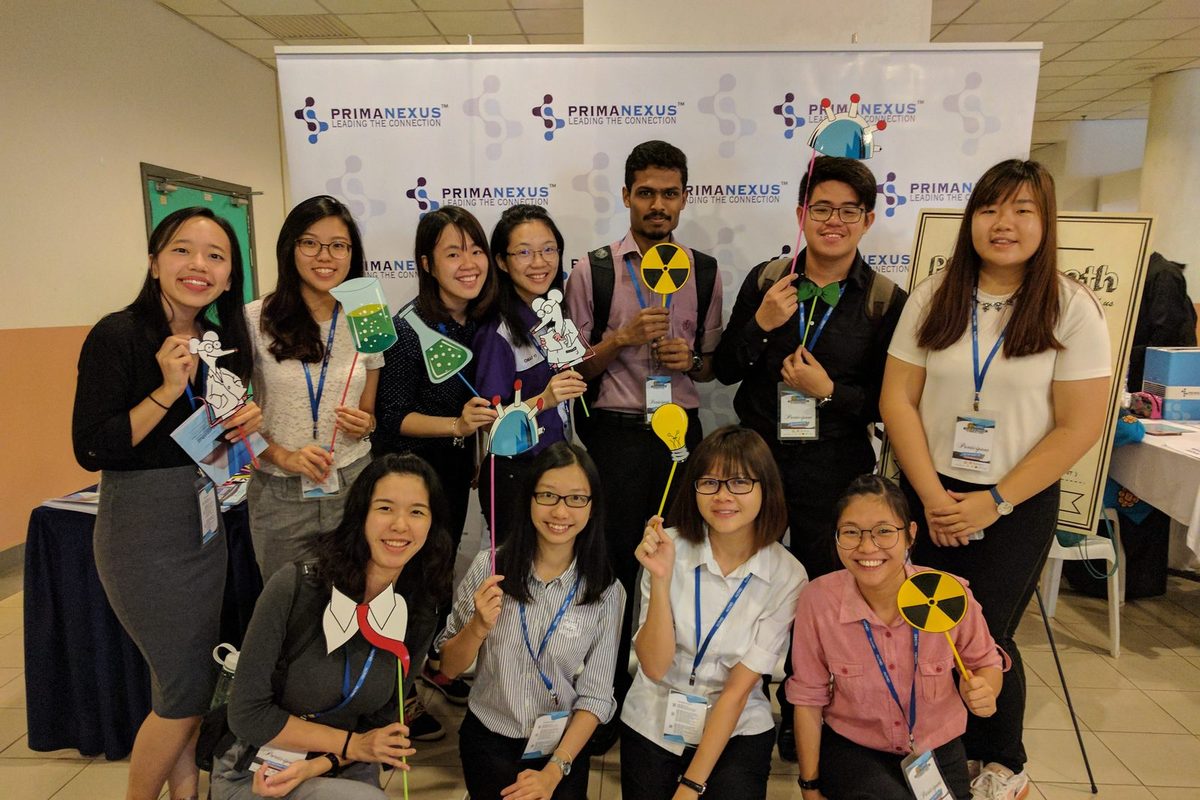 Smiling participants hold science-themed photo props in front of a Prima Nexus backdrop.