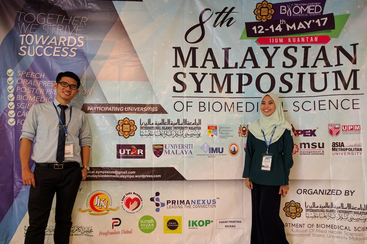 Two people stand beside the symposium banner, which carries the Prima Nexus logo.