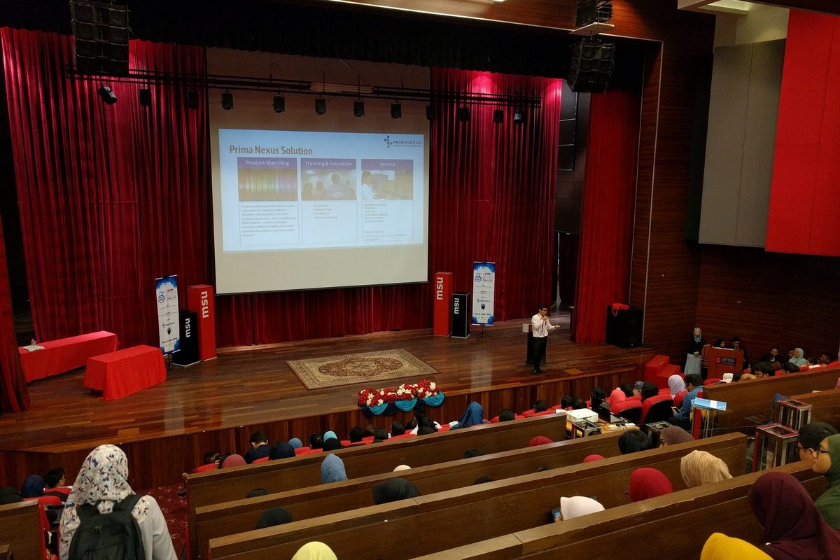 A speaker on the theatre stage with a Prima Nexus slide on the screen, and students seated in rows