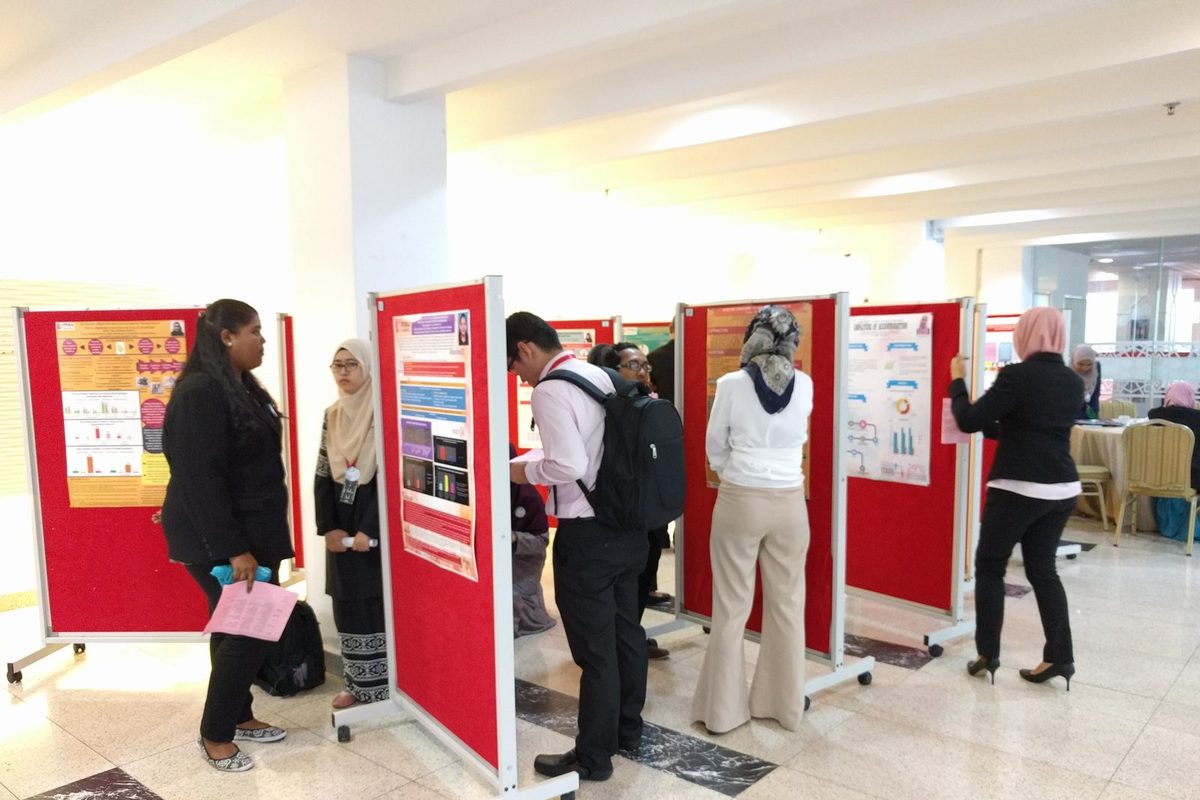 Students in formal dress reading research posters on red display boards in the foyer