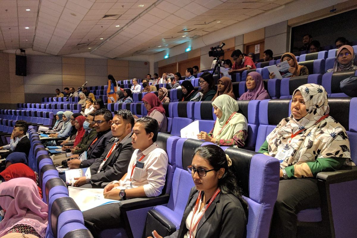 Delegates seated in rows of purple seats in a lecture hall, following a conference session