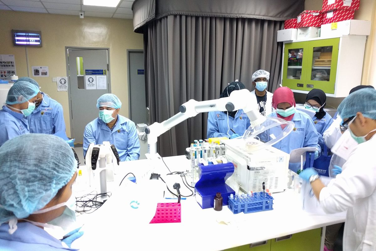 Participants in lab coats, masks and hair covers gather around a laboratory bench with pipettes and sample racks
