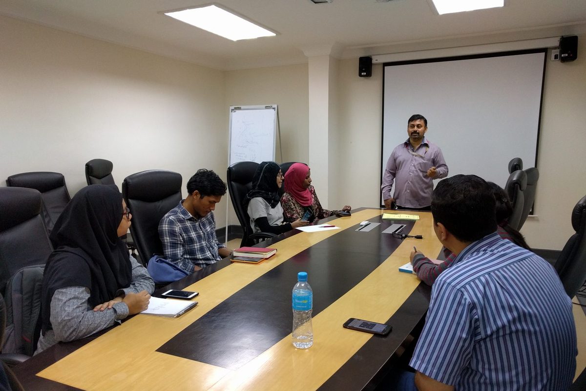 A speaker stands at the front of a meeting room while participants sit around the table