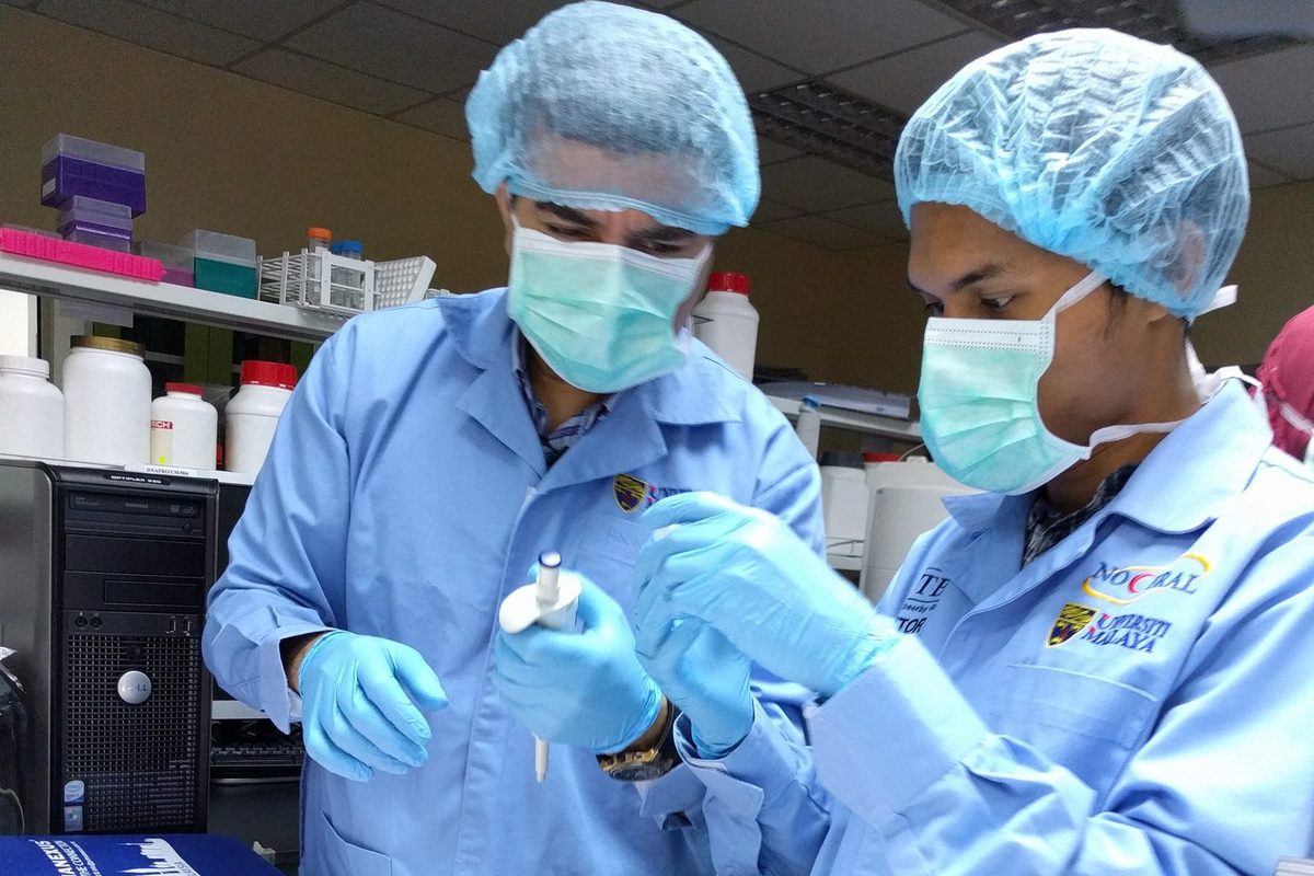 Two participants in hair covers and masks handle a pipette and sample tubes at the bench