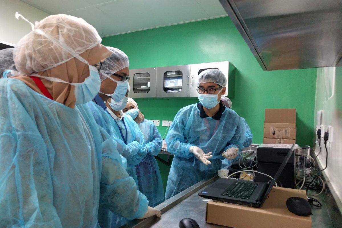 A trainer in gown and mask demonstrates the CODA system at a lab bench to a group of gowned participants