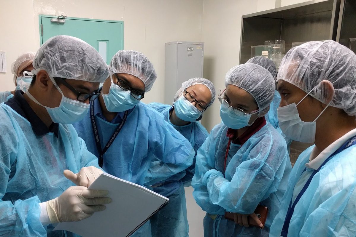 A trainer holds a spiral-bound manual while five gowned participants stand around and listen