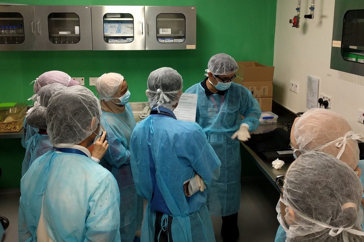 Participants in gowns and hair covers, seen from behind, gather around a trainer in the facility room