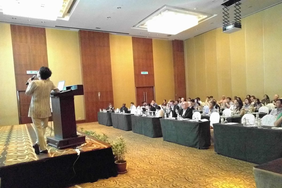 A speaker at the lectern addresses delegates seated at long tables in the conference hall