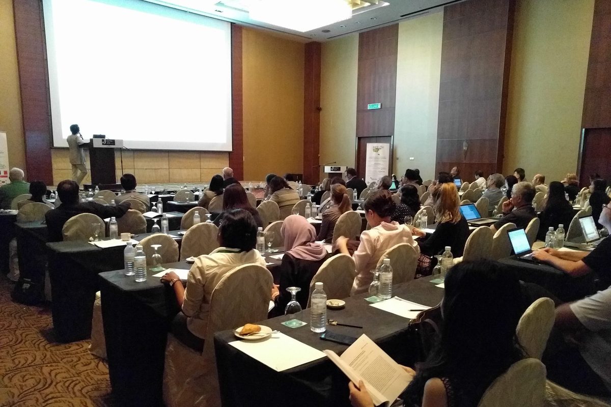 Delegates at long tables follow a talk, with the speaker and projection screen at the front of the hall