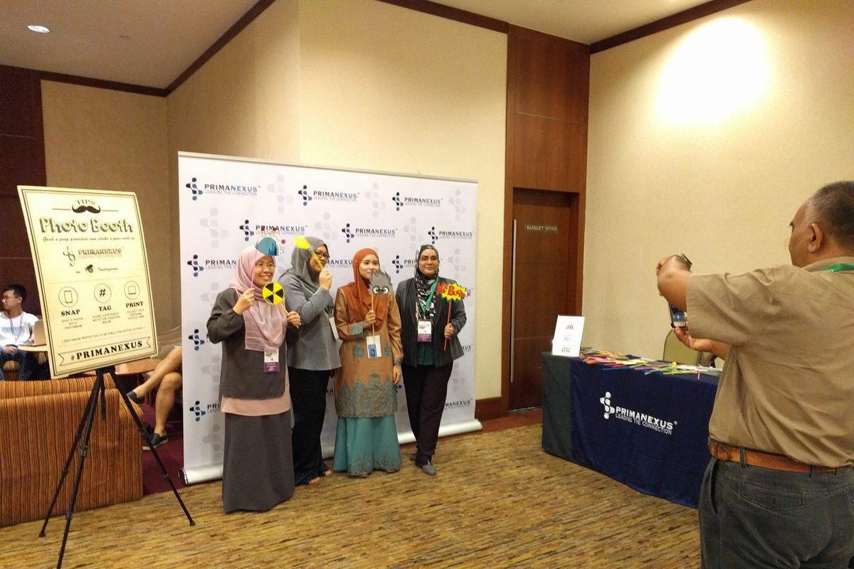 Four delegates hold photo props in front of the Prima Nexus backdrop while a man takes their picture