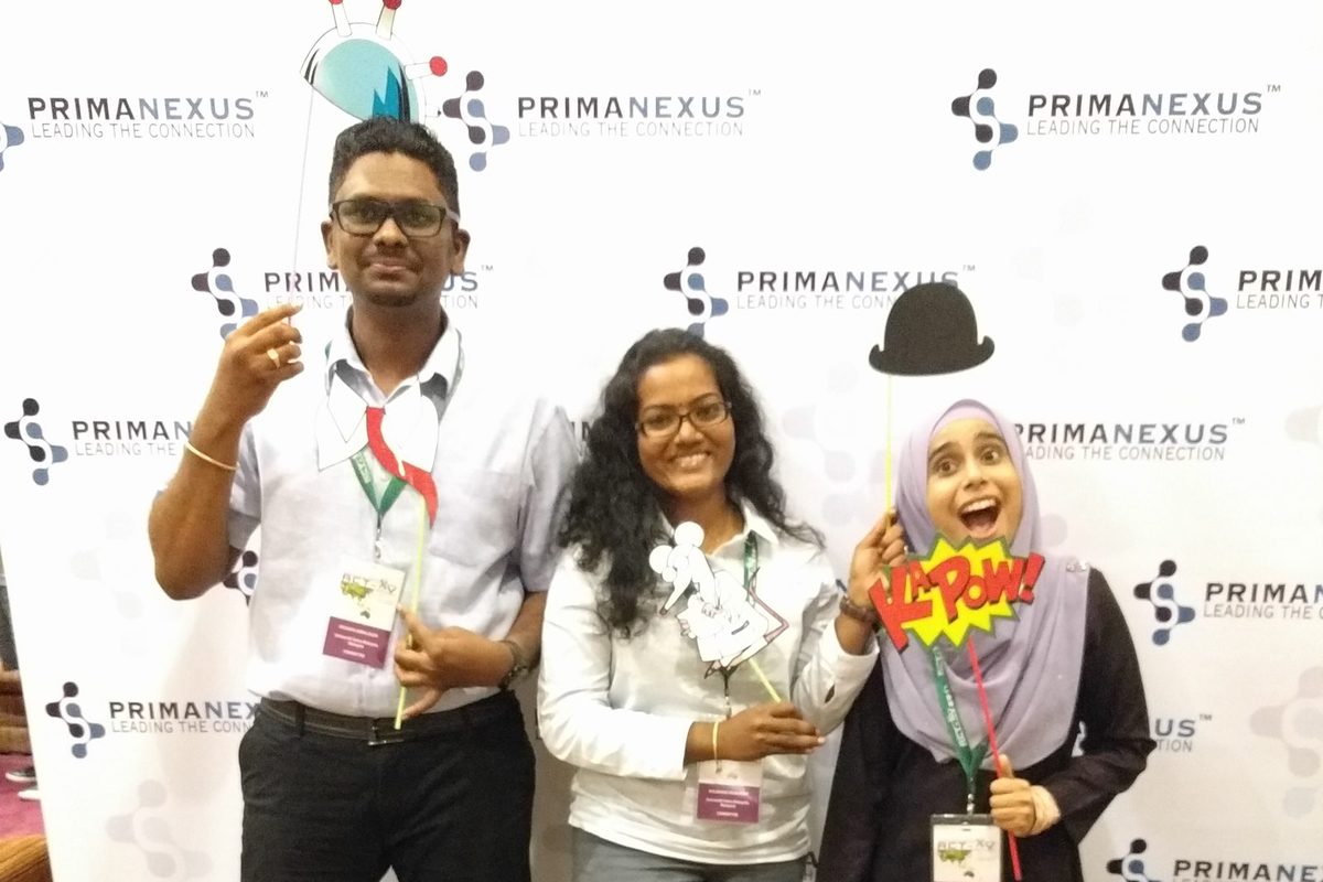 Three delegates with conference badges pose with props in front of the Prima Nexus backdrop