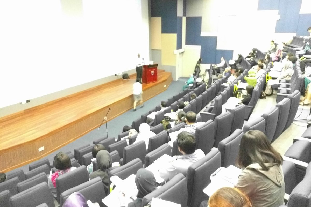 Delegates seated in the tiered auditorium while a lecturer stands at the podium on the stage