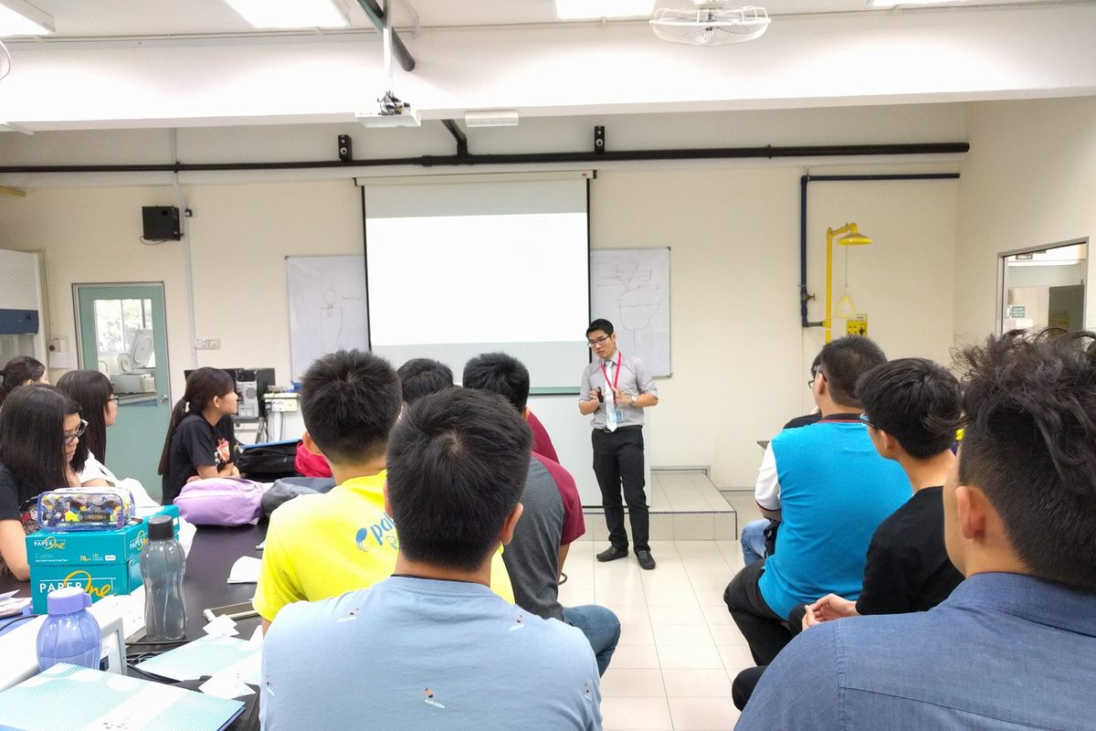 A speaker presents in front of a projection screen while participants listen from their benches