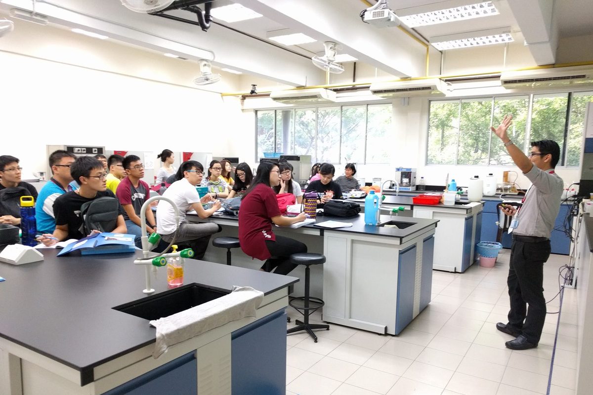 A presenter raises one hand while speaking to participants seated at laboratory benches