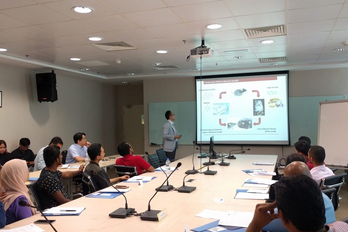 A presenter stands beside a projected slide at the front of the room, with researchers seated at long tables