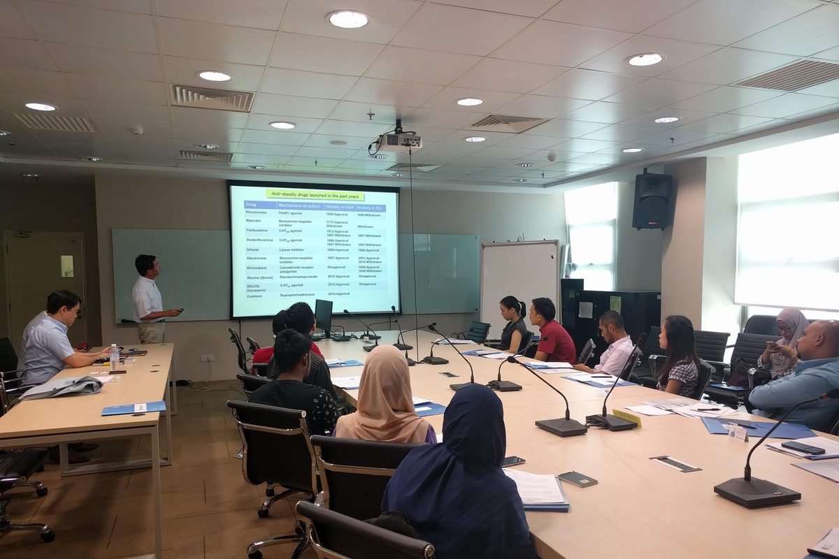A speaker at the front of the seminar room stands beside a projected table, with participants seated at long tables