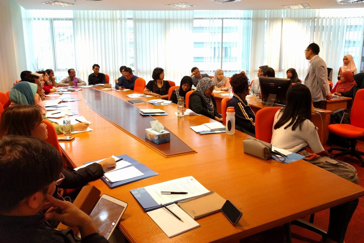 Participants seated around a long boardroom table while a speaker stands at the front of the room