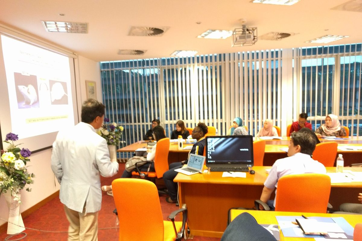 A speaker in a light jacket presents a slide showing laboratory rats to participants seated at desks.