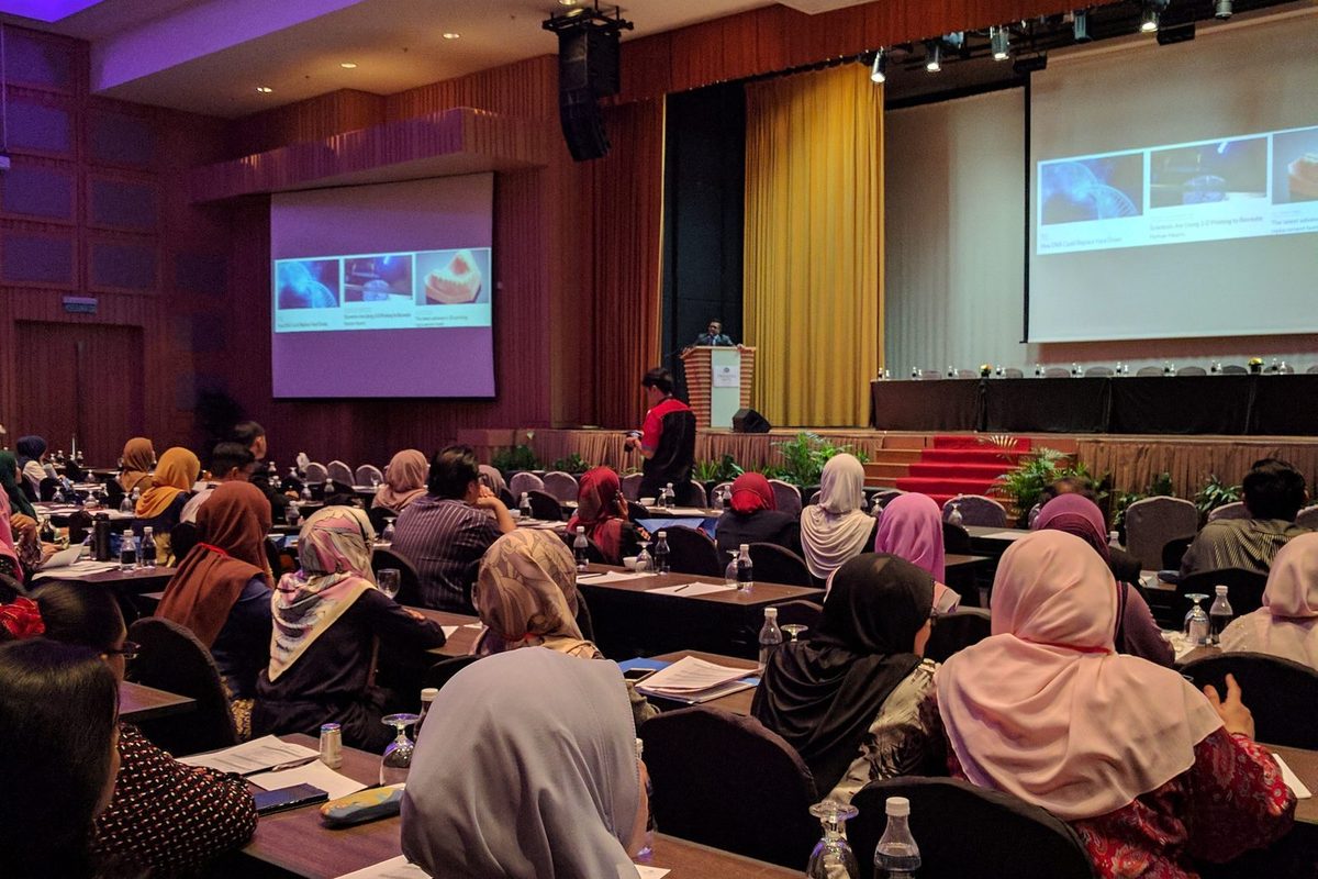 Delegates at tables listen to a talk, with slides shown on large screens beside the stage
