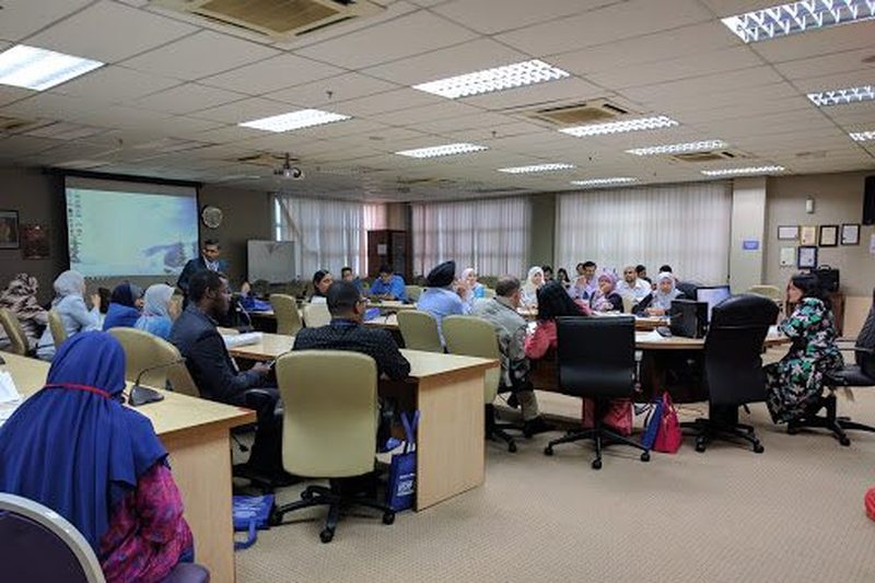 Delegates seated at desks and office chairs in a meeting room, with a projector screen at the front.