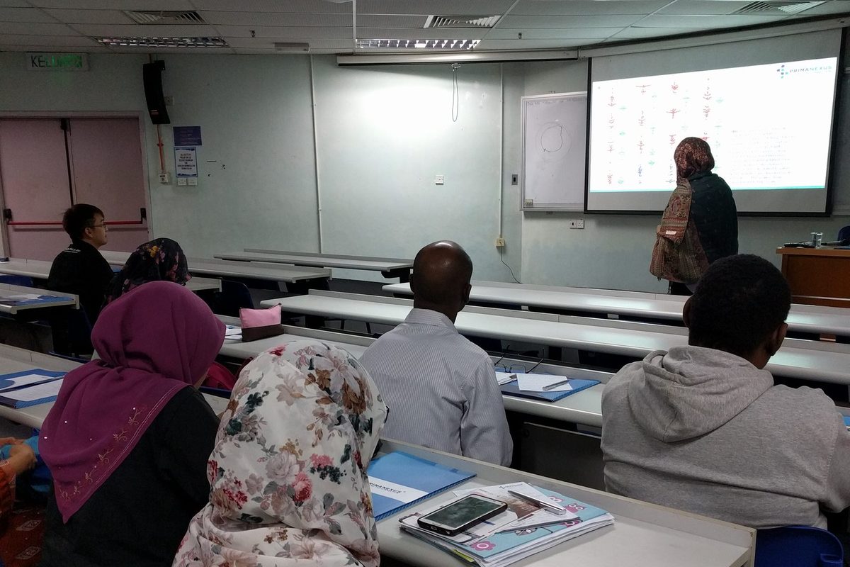 A presenter stands beside a projected slide while participants listen from their desks