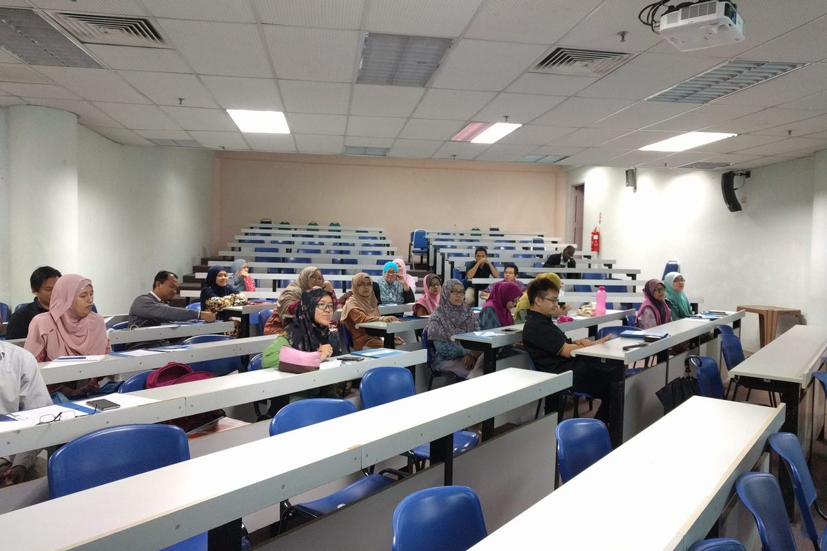 Participants seated at long desks in a tiered lecture room, facing the front