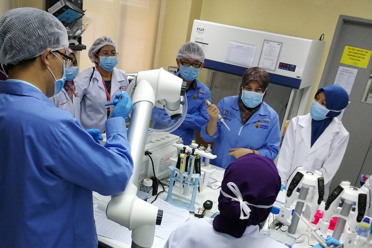 Participants in blue gowns pipette at a bench with the trainer