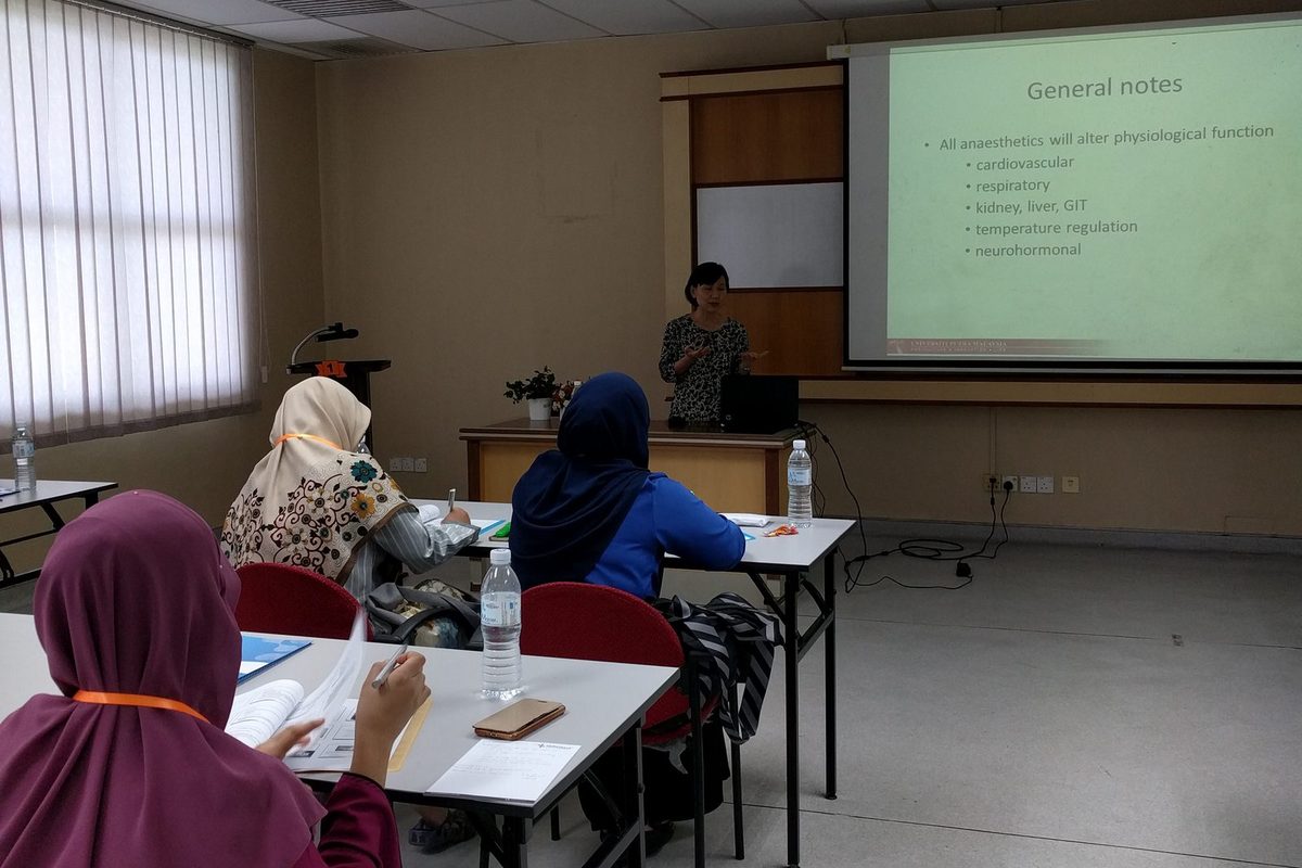 A lecture room with participants at tables facing a speaker and a slide on how anaesthetics affect the body