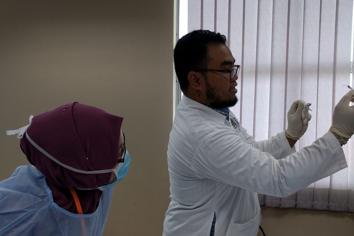 A man in a white lab coat holds up a syringe while a participant in a gown and mask watches
