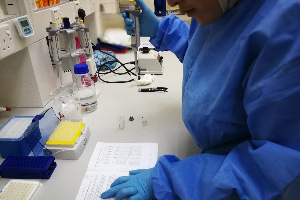 Gloved hands hold a pipette over a kit protocol at a lab bench, beside tube racks, reagent vials and a well plate.