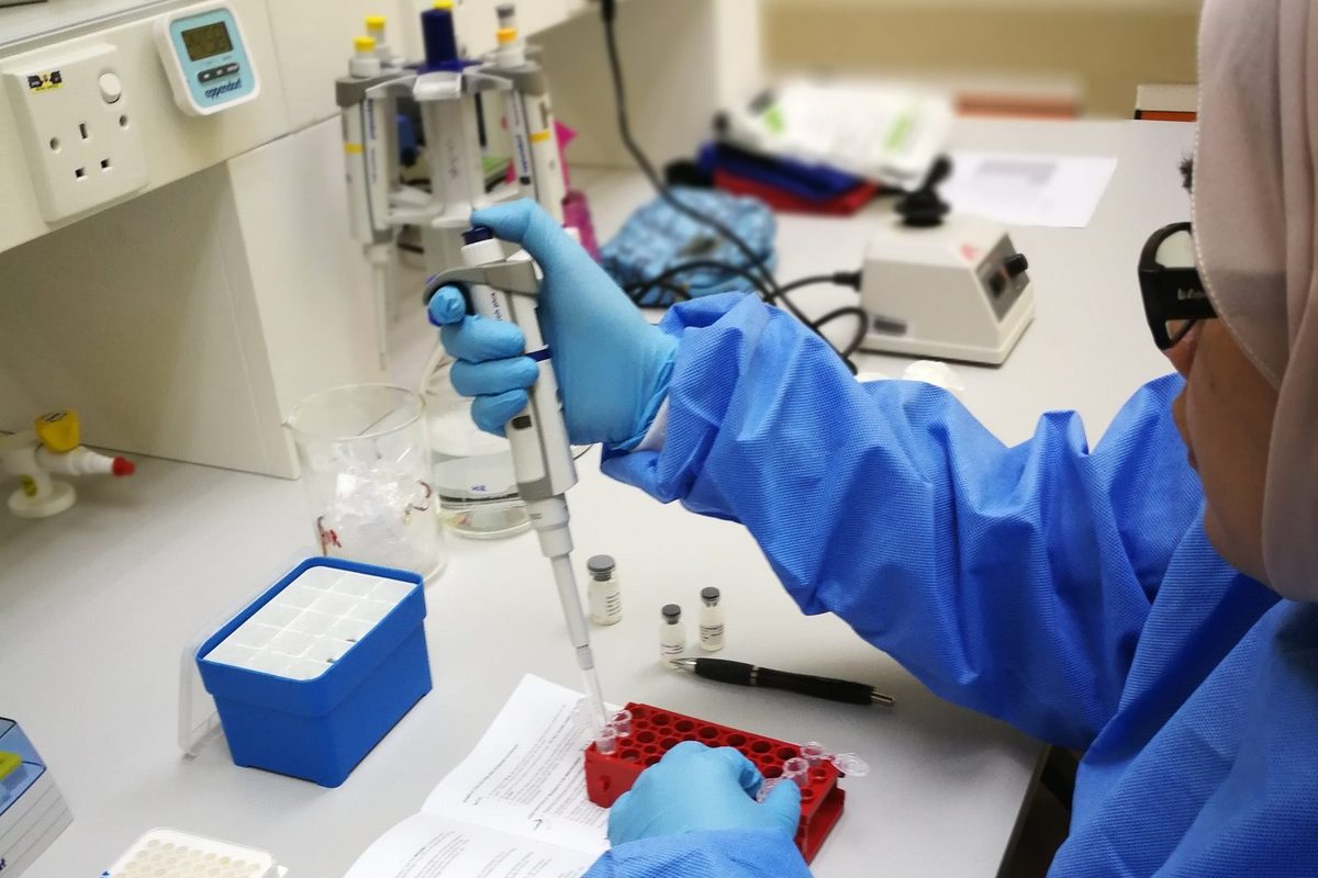 A researcher in a blue lab gown and gloves pipettes into a tube in a red rack, with the kit protocol open on the bench.