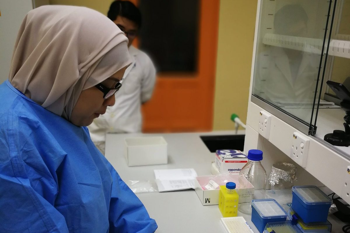 A researcher in a blue lab gown reads a kit insert at the bench while a man in a white coat looks on behind her.