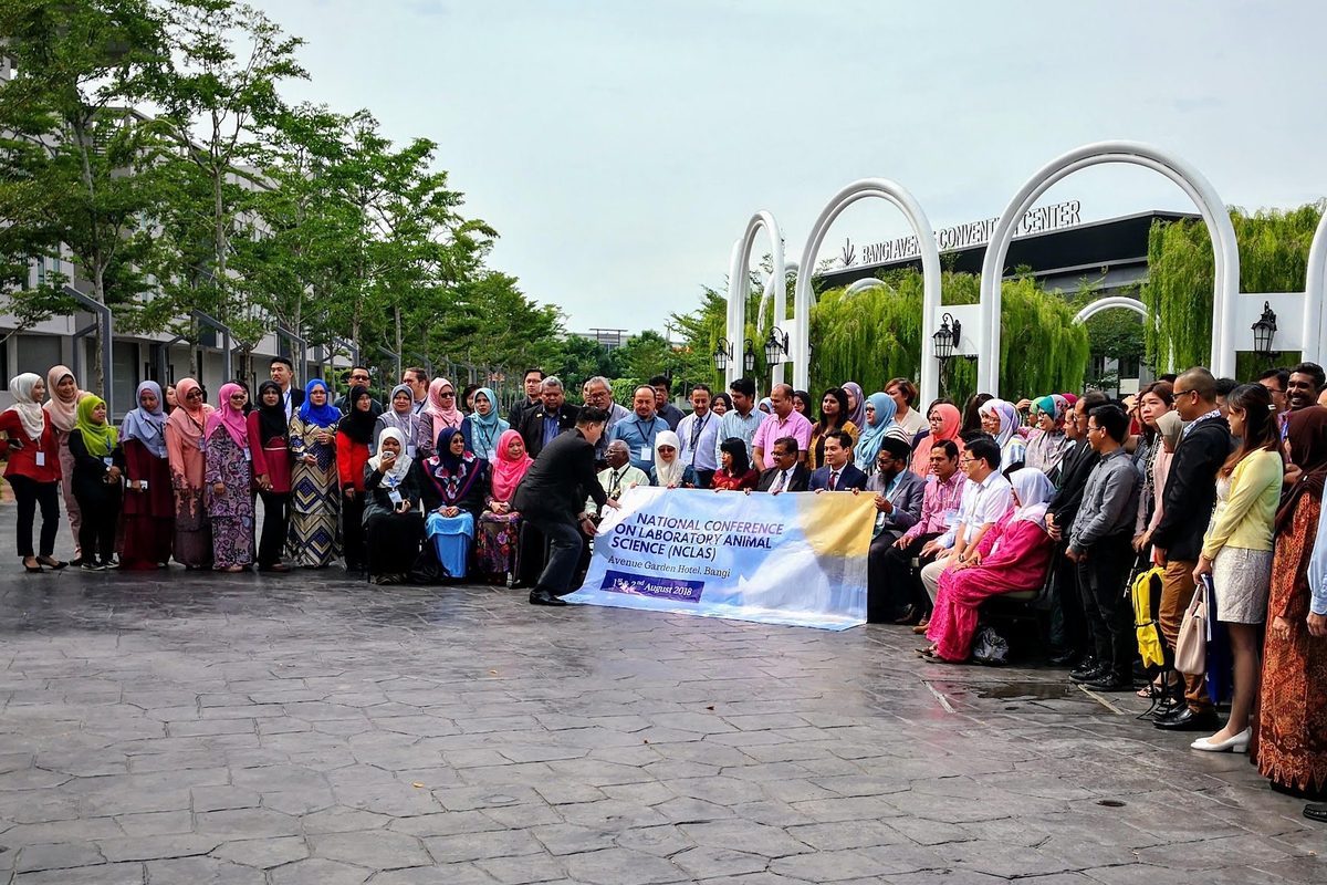 Conference participants in a large group photo outdoors in front of white arches, holding the conference banner