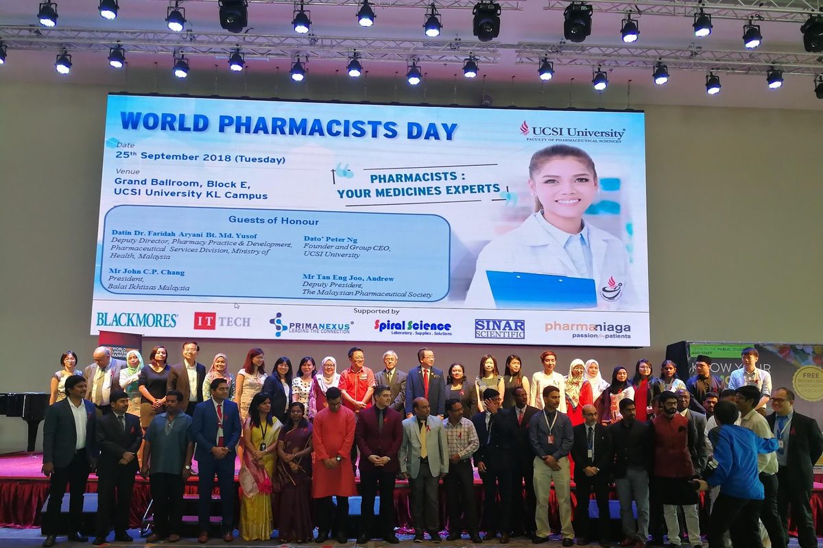 Students, staff and guests in a group photo on stage below the World Pharmacists Day screen