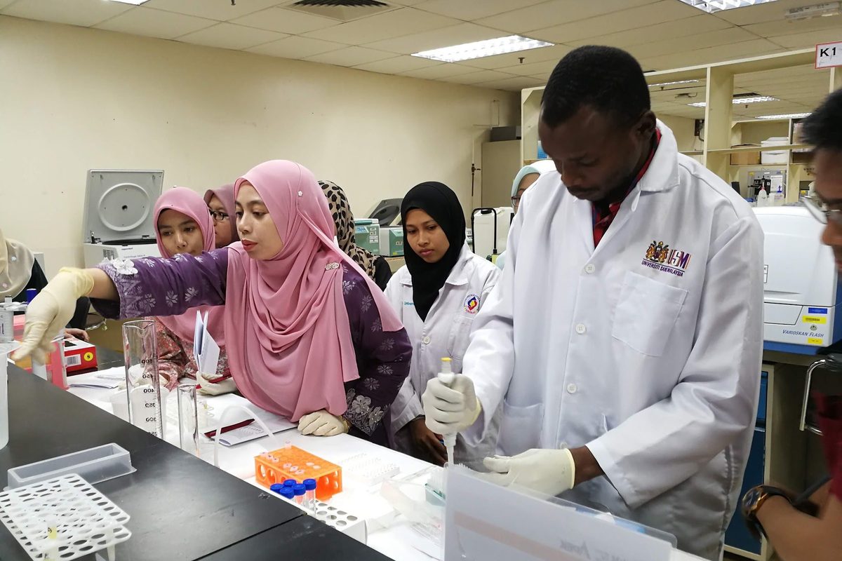 A trainer in a lab coat demonstrates at the bench while participants watch