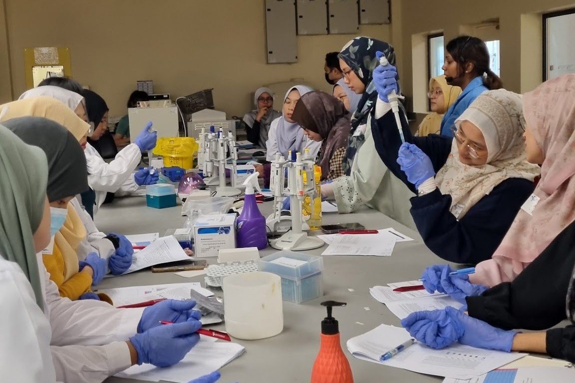 Participants in lab coats and gloves at long laboratory benches, one raising a hand