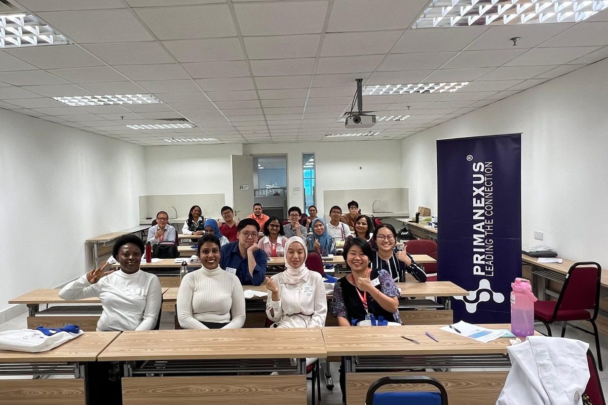 Workshop participants seated at tables in a classroom for a group photo, beside a Prima Nexus banner