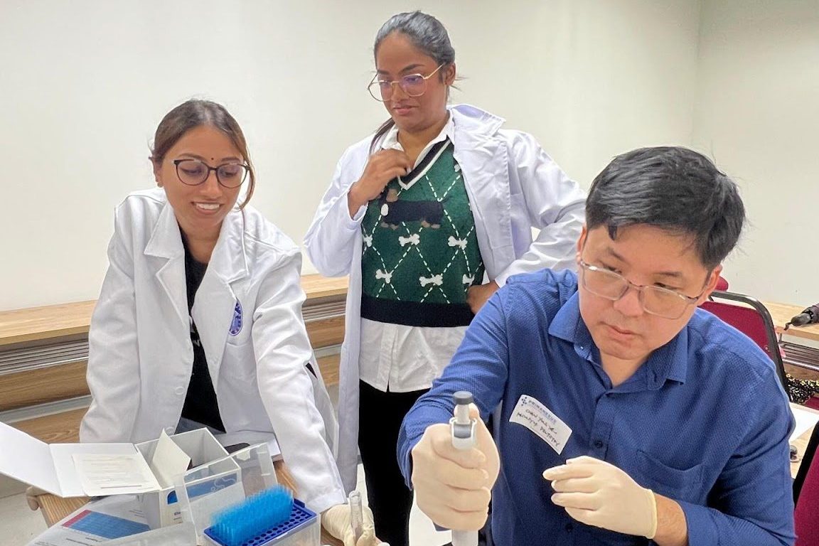 A trainer in a lab coat works with participants at a table of workshop materials
