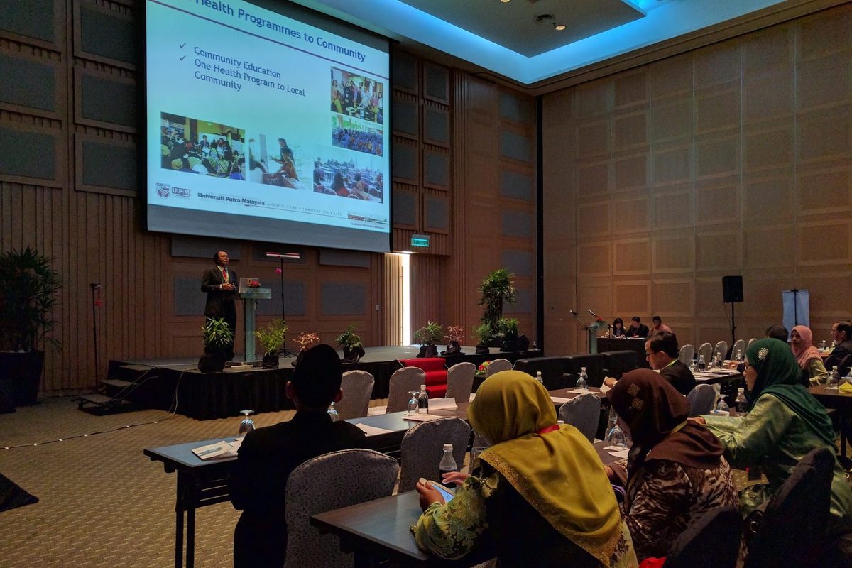 A speaker presents to delegates seated at long tables in a conference hall, with slides on a large screen